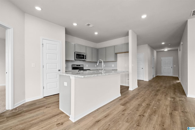 kitchen with gray cabinetry, light stone counters, appliances with stainless steel finishes, an island with sink, and light hardwood / wood-style floors