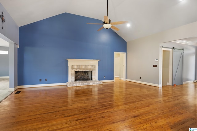 unfurnished living room with ceiling fan, a barn door, high vaulted ceiling, light wood-type flooring, and a fireplace