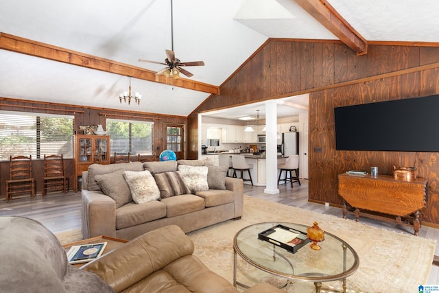 living room with ceiling fan with notable chandelier, vaulted ceiling with beams, wood walls, and light hardwood / wood-style flooring