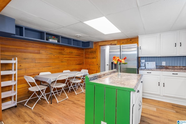 kitchen featuring light wood-type flooring, wooden walls, stainless steel fridge, white cabinetry, and a center island with sink