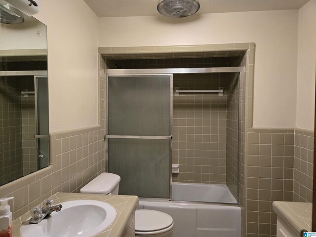 bathroom with tile walls, a sink, toilet, shower / bath combination with glass door, and a wainscoted wall