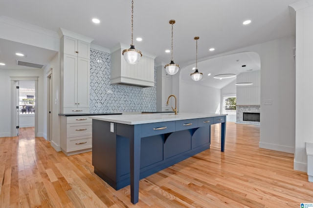 kitchen with light hardwood / wood-style flooring, pendant lighting, a kitchen island with sink, a fireplace, and white cabinets