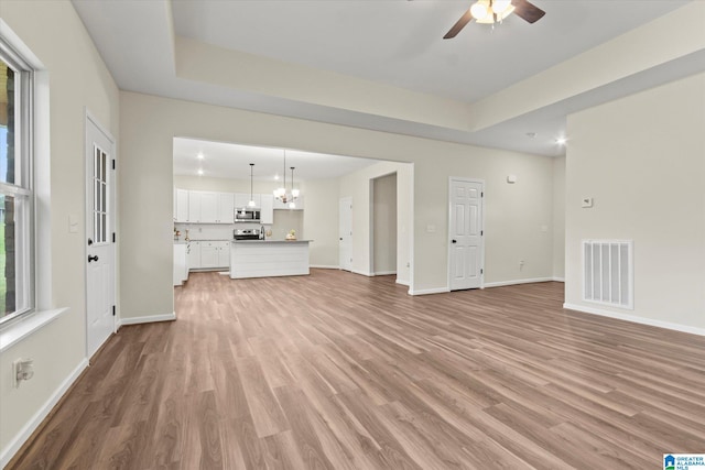 unfurnished living room with ceiling fan with notable chandelier, light wood-type flooring, and a raised ceiling