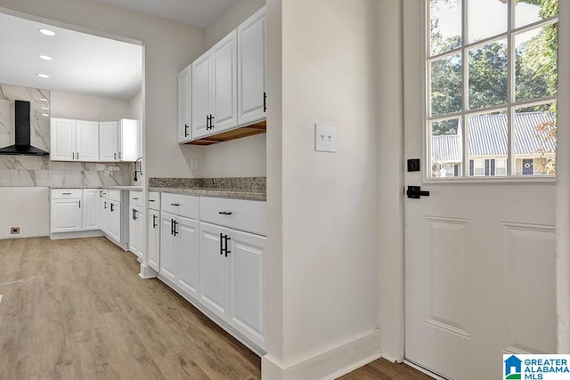 kitchen featuring wall chimney range hood, light hardwood / wood-style flooring, white cabinetry, and decorative backsplash