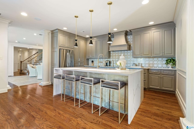 kitchen with stainless steel fridge, custom range hood, dark wood-type flooring, and an island with sink