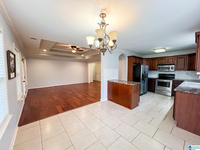 kitchen with ornamental molding, light wood-type flooring, appliances with stainless steel finishes, a raised ceiling, and ceiling fan with notable chandelier