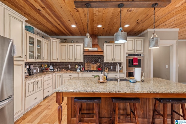 kitchen with a kitchen island with sink, cream cabinets, light hardwood / wood-style flooring, decorative light fixtures, and light stone counters