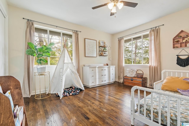 bedroom featuring multiple windows, a nursery area, dark wood-type flooring, and ceiling fan