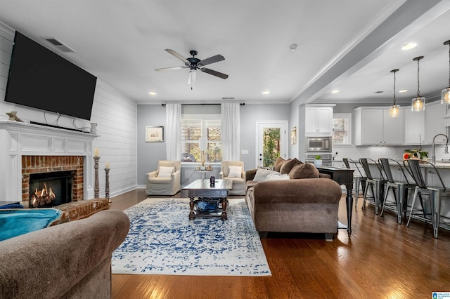 living room featuring a brick fireplace, ceiling fan, wooden walls, dark wood-type flooring, and ornamental molding