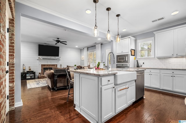 kitchen featuring white cabinetry, a kitchen island with sink, a fireplace, decorative light fixtures, and light stone counters