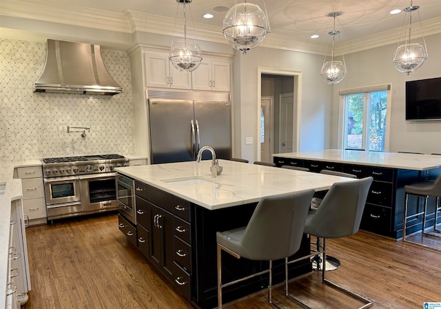 kitchen with white cabinets, a sink, wall chimney range hood, an island with sink, and built in appliances