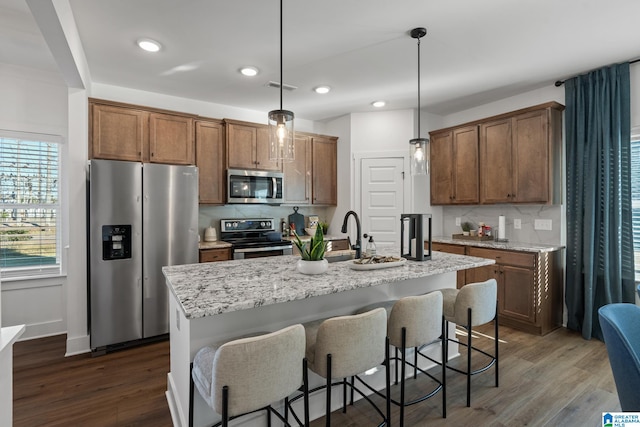 kitchen with hanging light fixtures, stainless steel appliances, a center island with sink, light stone countertops, and dark hardwood / wood-style flooring