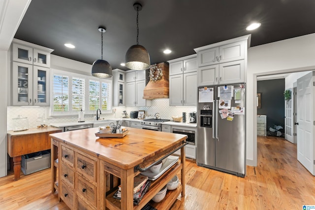 kitchen featuring decorative light fixtures, light wood-type flooring, custom exhaust hood, appliances with stainless steel finishes, and tasteful backsplash