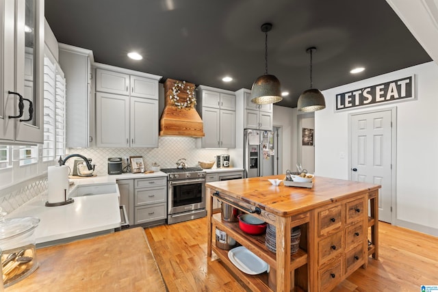 kitchen with gray cabinetry, appliances with stainless steel finishes, hanging light fixtures, light hardwood / wood-style floors, and custom range hood