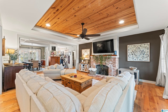 living room featuring ceiling fan with notable chandelier, a tray ceiling, light wood-type flooring, and a brick fireplace