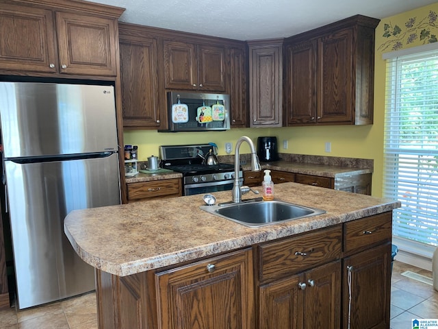 kitchen with an island with sink, dark brown cabinets, stainless steel appliances, sink, and light tile patterned floors