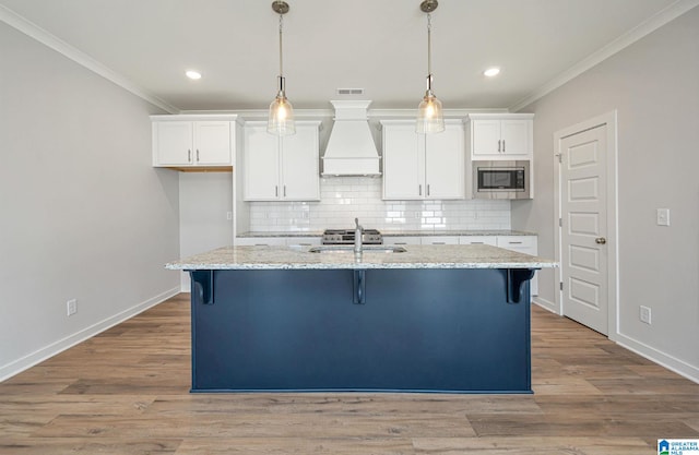 kitchen with light stone counters, stainless steel microwave, premium range hood, a center island with sink, and white cabinetry