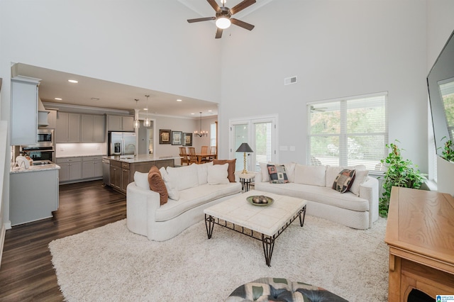 living room featuring dark wood-type flooring, a healthy amount of sunlight, a high ceiling, and ceiling fan with notable chandelier