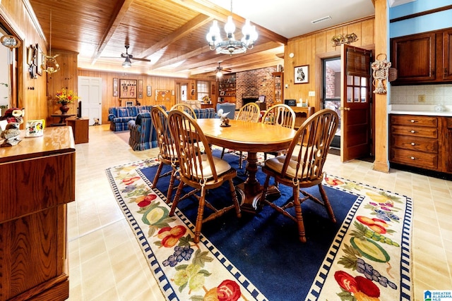 tiled dining room featuring a brick fireplace, ceiling fan with notable chandelier, wooden walls, and beam ceiling