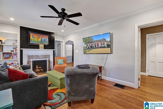 living room featuring built in shelves, ceiling fan, a large fireplace, hardwood / wood-style floors, and crown molding