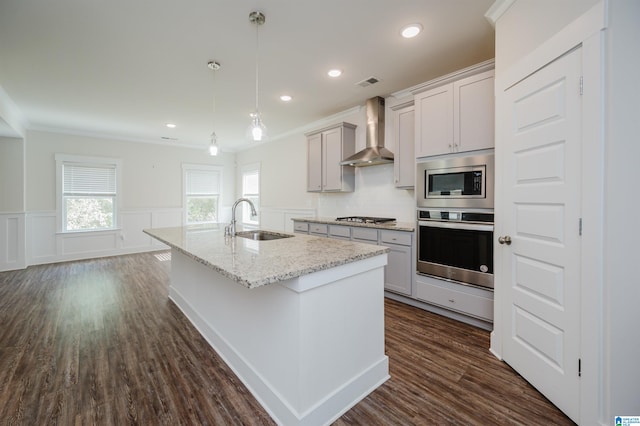 kitchen featuring stainless steel appliances, wall chimney range hood, decorative light fixtures, sink, and a kitchen island with sink