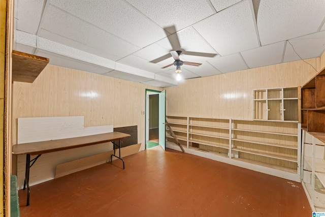 basement featuring a paneled ceiling, wood walls, and ceiling fan