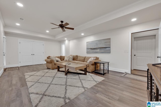 living room with ceiling fan, a raised ceiling, light wood-type flooring, and crown molding