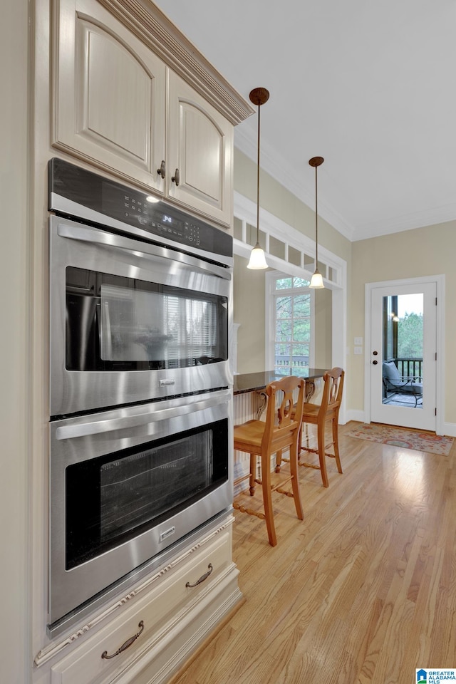 kitchen with light hardwood / wood-style floors, decorative light fixtures, stainless steel double oven, crown molding, and cream cabinetry