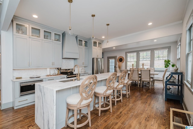 kitchen featuring a center island with sink, light stone countertops, appliances with stainless steel finishes, decorative light fixtures, and premium range hood