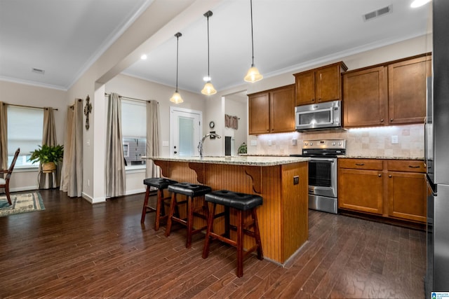 kitchen with appliances with stainless steel finishes, light stone counters, decorative light fixtures, dark hardwood / wood-style floors, and an island with sink