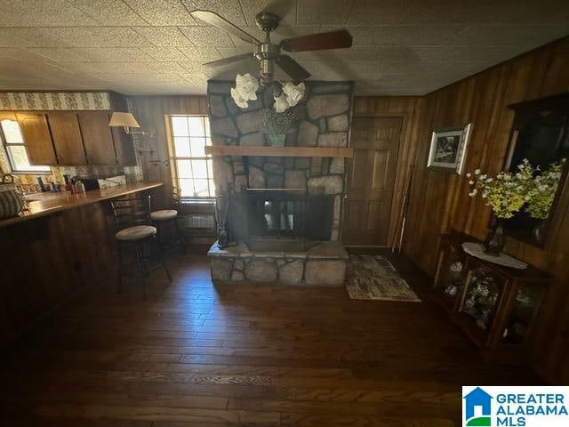 living room with a stone fireplace, ceiling fan, wooden walls, and dark hardwood / wood-style floors