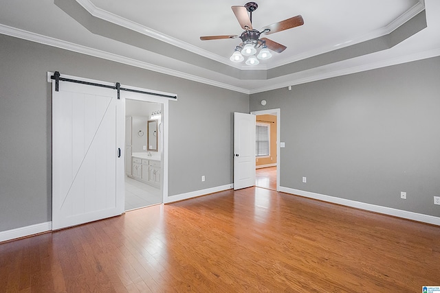 unfurnished bedroom with a barn door, ornamental molding, connected bathroom, a tray ceiling, and light hardwood / wood-style floors