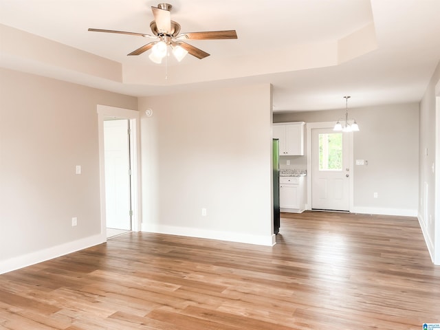 unfurnished living room with ceiling fan with notable chandelier, a raised ceiling, and light wood-type flooring