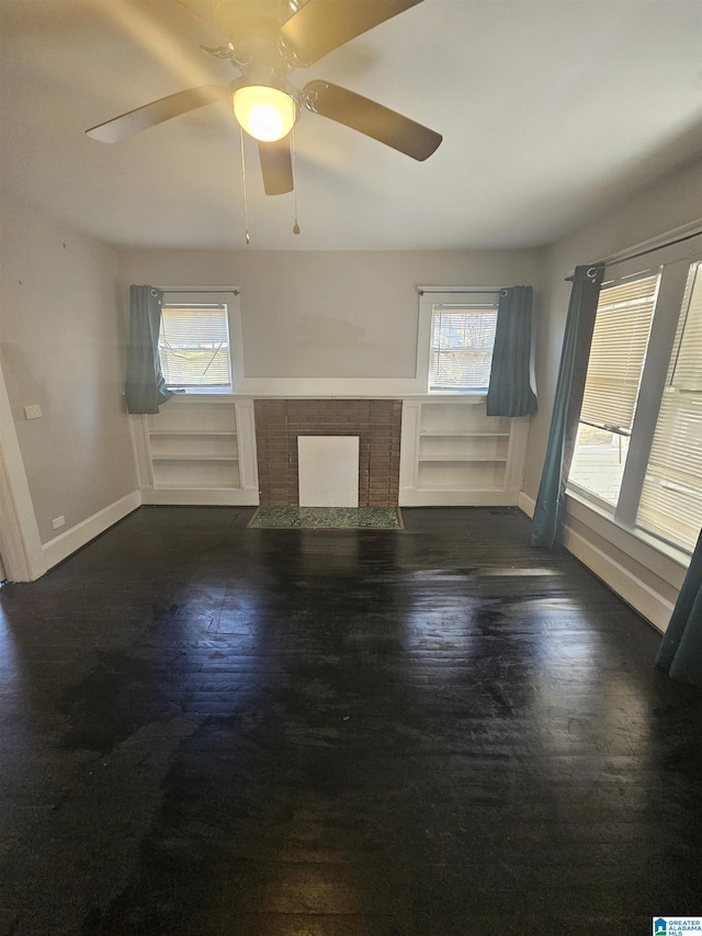 unfurnished living room with built in shelves, ceiling fan, dark wood-type flooring, and a brick fireplace