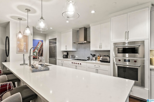 kitchen featuring wall chimney exhaust hood, stainless steel double oven, pendant lighting, a center island with sink, and white cabinetry