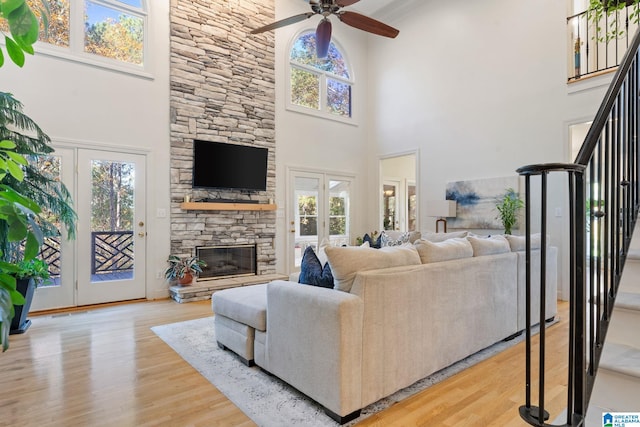 living room featuring ceiling fan, french doors, a stone fireplace, a towering ceiling, and light wood-type flooring