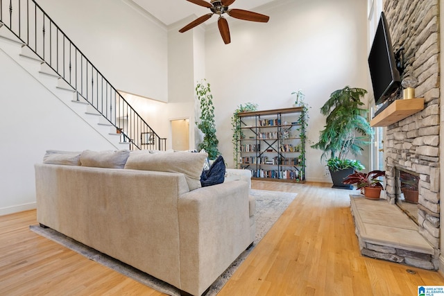 living room featuring a towering ceiling, light wood-type flooring, ornamental molding, ceiling fan, and a stone fireplace