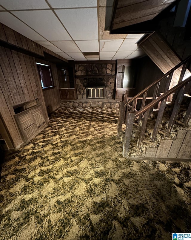 interior space featuring a drop ceiling, wood walls, a stone fireplace, and carpet floors