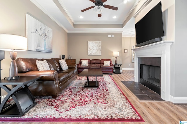 living room featuring ceiling fan, crown molding, wood-type flooring, a tray ceiling, and a tiled fireplace