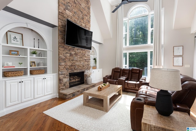 living room with built in shelves, ceiling fan, wood-type flooring, high vaulted ceiling, and a stone fireplace