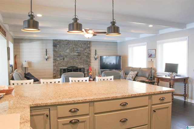 kitchen with beam ceiling, ceiling fan, a stone fireplace, light stone counters, and dark hardwood / wood-style floors