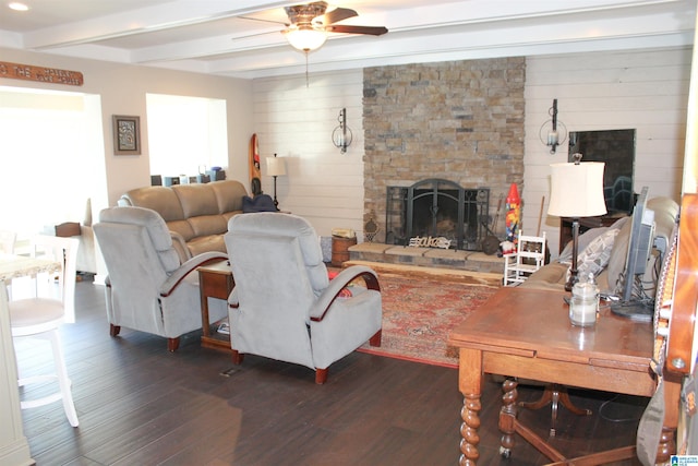 living room with beam ceiling, a stone fireplace, ceiling fan, and dark wood-type flooring