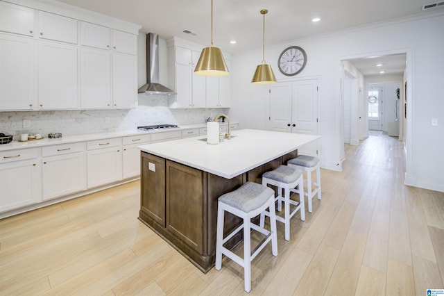 kitchen with sink, wall chimney exhaust hood, an island with sink, pendant lighting, and white cabinets