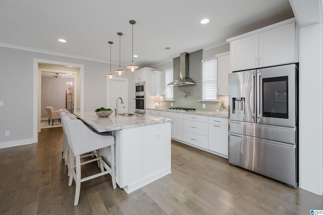 kitchen featuring wall chimney range hood, hardwood / wood-style flooring, an island with sink, appliances with stainless steel finishes, and white cabinetry