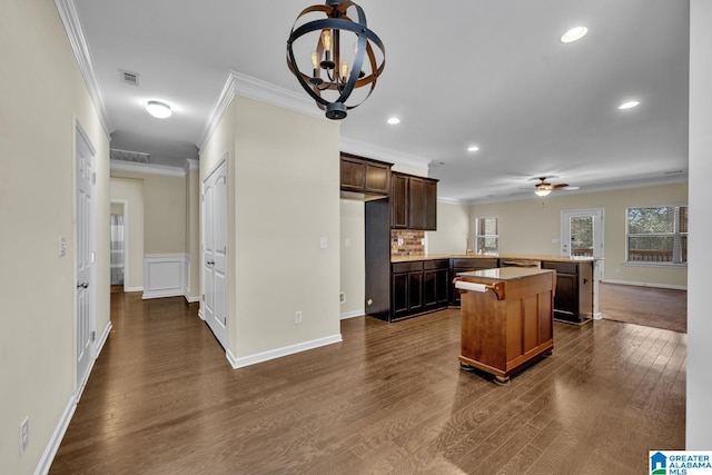 kitchen with dark wood-type flooring, crown molding, dark brown cabinets, a kitchen island, and ceiling fan with notable chandelier