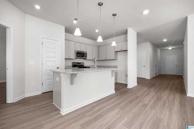 kitchen with pendant lighting, appliances with stainless steel finishes, light hardwood / wood-style floors, light stone countertops, and an island with sink