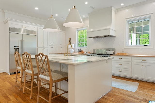 kitchen with custom exhaust hood, hanging light fixtures, a kitchen island with sink, and appliances with stainless steel finishes