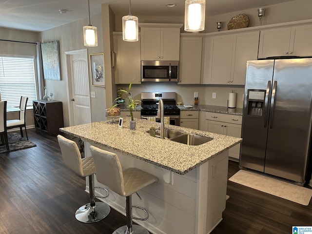 kitchen with dark wood-type flooring, light stone counters, an island with sink, decorative light fixtures, and appliances with stainless steel finishes