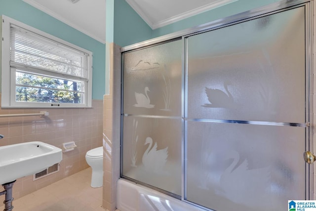 bathroom with tile patterned flooring, crown molding, combined bath / shower with glass door, toilet, and tile walls