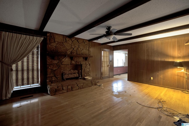 unfurnished living room featuring ceiling fan, a stone fireplace, beamed ceiling, wood-type flooring, and a textured ceiling